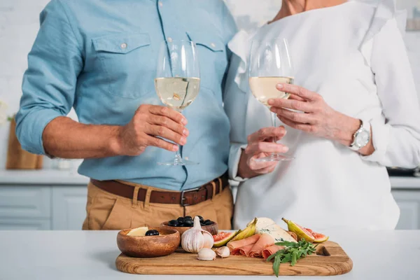Tiro recortado si pareja de ancianos sosteniendo vasos de vino mientras come deliciosos bocadillos en casa - foto de stock