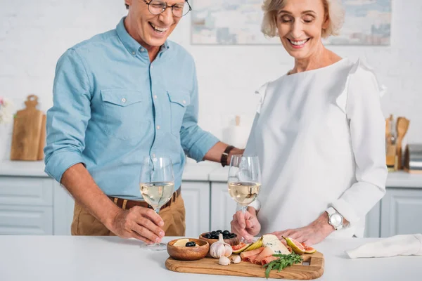 Cropped shot of smiling senior couple holding glasses of wine and looking at delicious snacks at home — Stock Photo