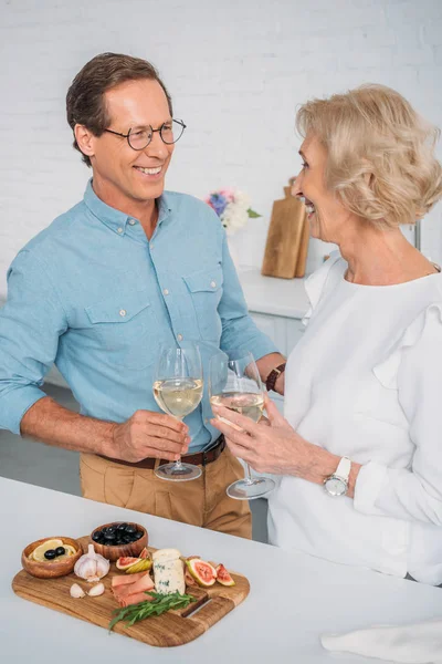 Feliz pareja de ancianos sosteniendo vasos de vino y sonriéndose mientras comen deliciosos bocadillos en casa - foto de stock