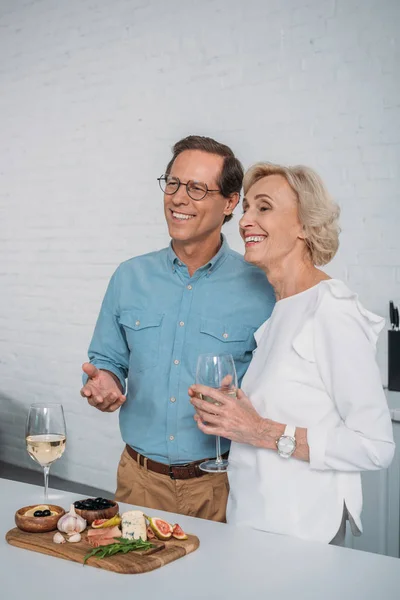 Feliz pareja de ancianos bebiendo vino y mirando hacia otro lado mientras come deliciosos bocadillos en casa - foto de stock