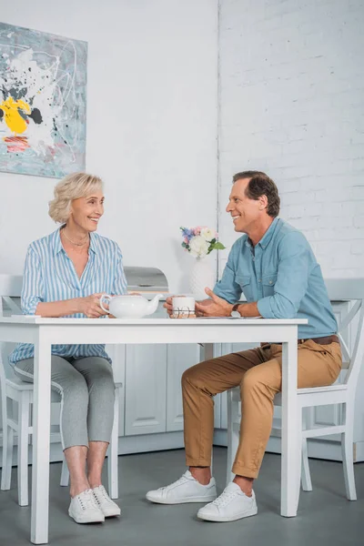 Feliz casal sênior sorrindo uns aos outros enquanto bebem chá juntos em casa — Fotografia de Stock