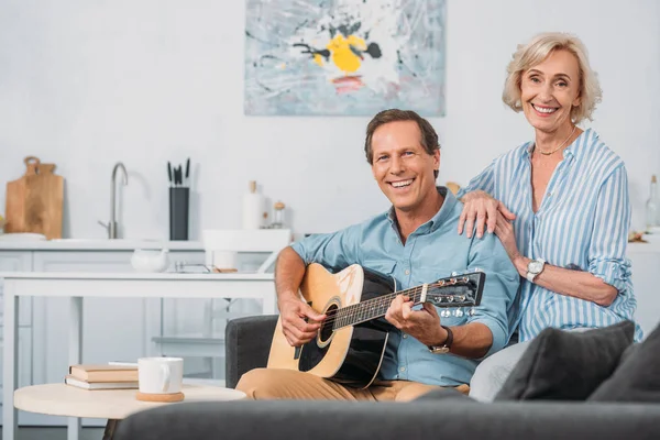 Heureux couple de personnes âgées souriant à la caméra tandis que l'homme jouer de la guitare à la maison — Photo de stock