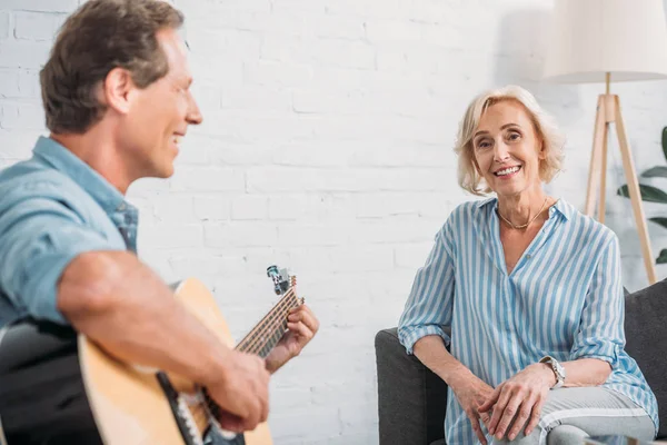 Счастливый мужчина играет на гитаре с улыбчивой старшей женой дома — стоковое фото