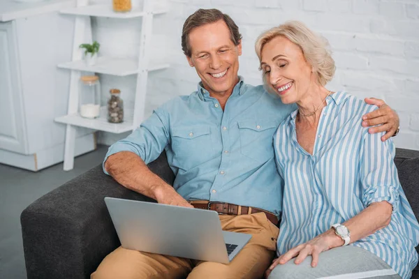 Sonriendo pareja de ancianos utilizando el ordenador portátil mientras están sentados juntos en casa - foto de stock