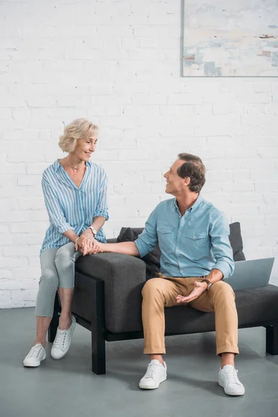 Feliz casal sênior de mãos dadas e sorrindo uns aos outros enquanto sentados juntos em casa — Fotografia de Stock