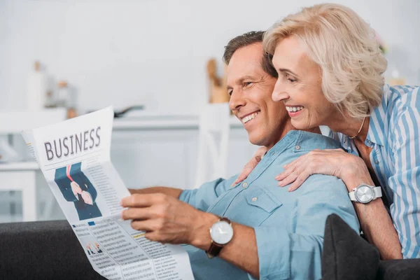 Vue latérale d'un couple âgé souriant lisant un journal d'affaires à la maison — Photo de stock