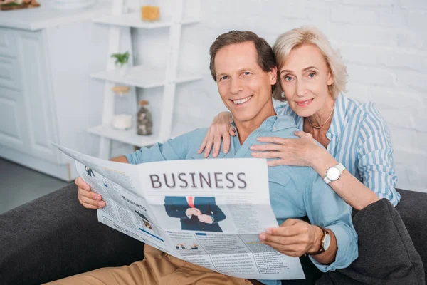 Glückliches Seniorenpaar lächelt in die Kamera, während es zu Hause die Wirtschaftszeitung liest — Stockfoto