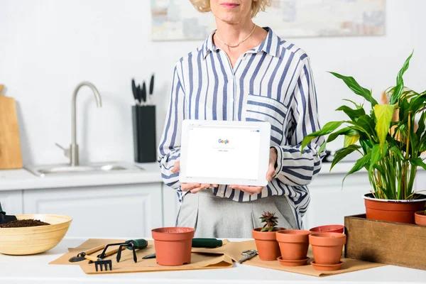 Tiro recortado de mulher sênior segurando tablet digital com página do google carregado enquanto cultivando plantas em casa — Fotografia de Stock