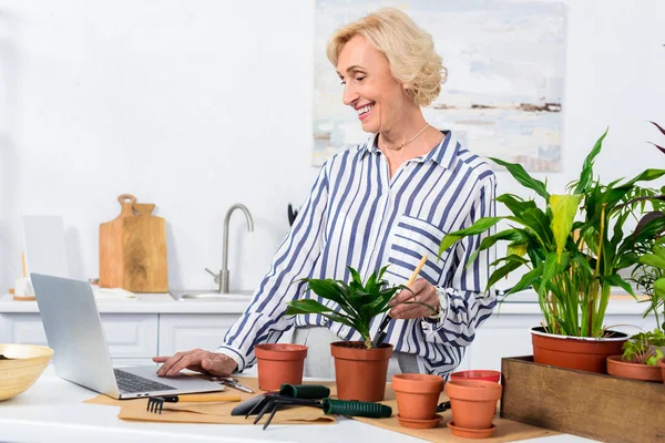 Sorrindo mulher idosa usando laptop e cultivando plantas em vaso em casa — Fotografia de Stock