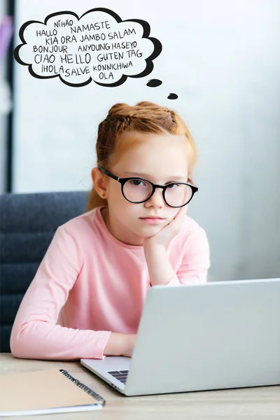 Aburrido niño pequeño en gafas mirando a la cámara mientras que srtudying con el ordenador portátil, con palabras en diferentes idiomas en burbuja del habla - foto de stock