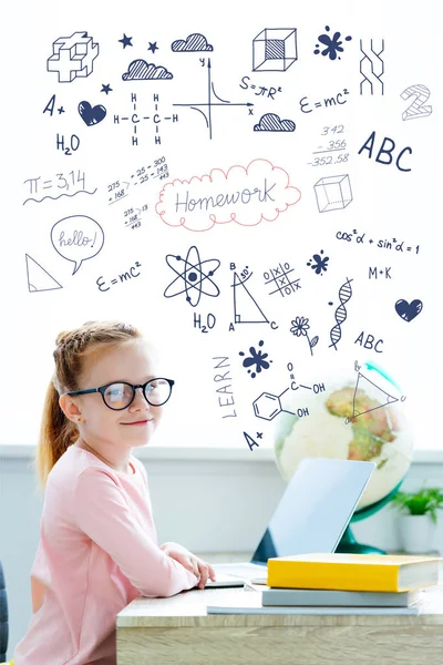 Belle écolière rousse aux lunettes souriant à la caméra tout en étudiant avec des livres et un ordinateur portable, avec des devoirs et des icônes éducatives — Photo de stock