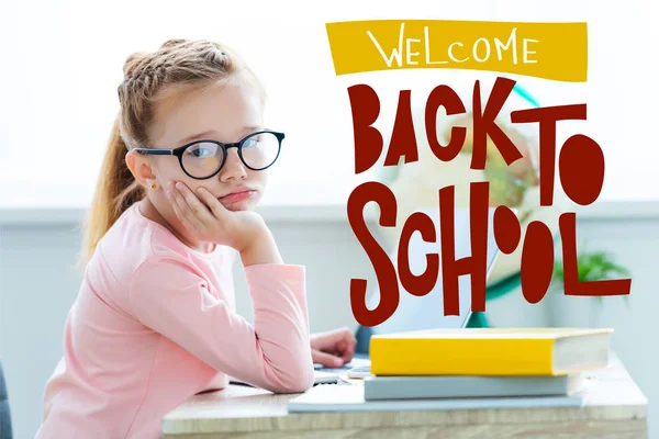 Bored child in eyeglasses looking at camera while studying with laptop and books, with 