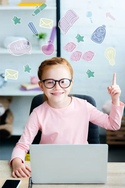 Adorable écolière rousse aux lunettes pointant vers le haut avec le doigt et souriant à la caméra tout en utilisant un ordinateur portable à la maison avec des icônes éducatives — Photo de stock