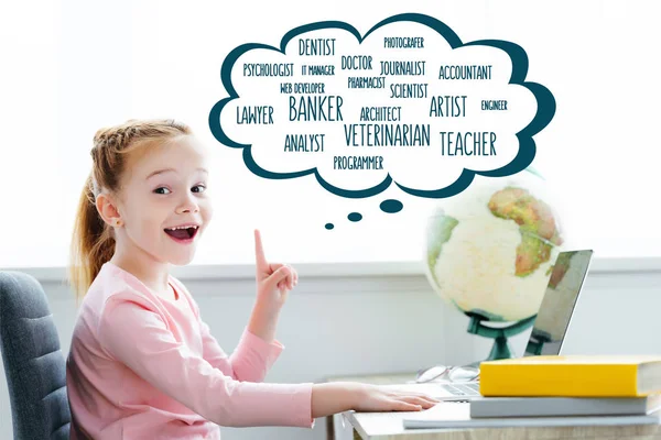 Cheerful redhead child studying with books and laptop and pointing up on words of different professions in speech bubble — Stock Photo