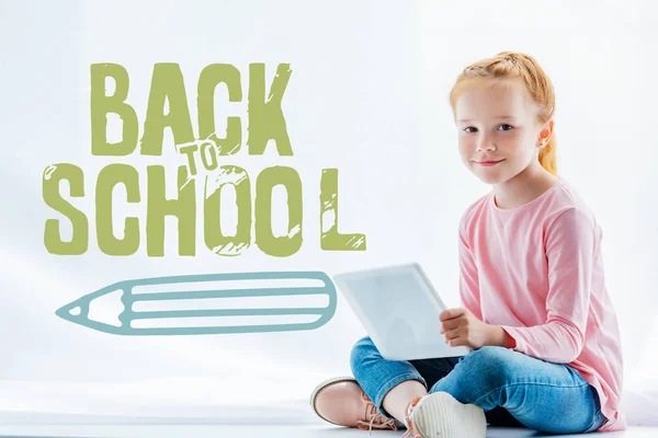 Hermosa pelirroja niño utilizando tableta digital y sonriendo a la cámara mientras está sentado en el alféizar de la ventana, de vuelta al concepto de la escuela - foto de stock