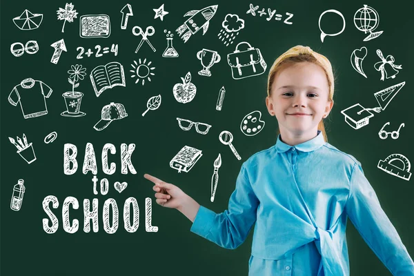 Belle écolière aux cheveux rouges souriants pointant vers le tableau noir avec des icônes et lettrage de retour à l'école — Photo de stock