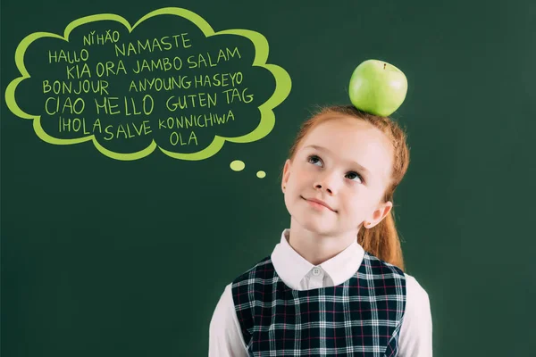 Pensativa colegiala pelirroja con manzana en la cabeza de pie cerca de pizarra con palabras en diferentes idiomas en la burbuja del habla - foto de stock