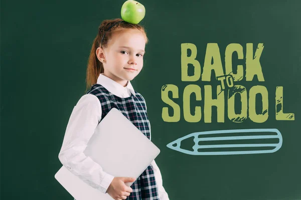 Belle petite écolière avec pomme sur la tête tenant ordinateur portable et debout près du tableau noir avec lettrage de retour à l'école — Photo de stock