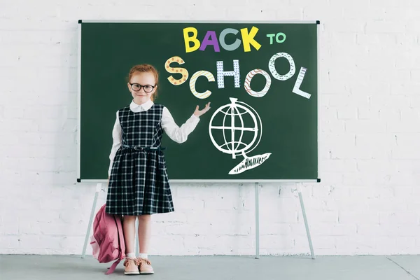 Adorable little schoolgirl with backpack showing blackboard with back to school lettering — Stock Photo