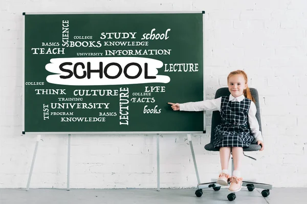 Adorable colegiala sonriendo a la cámara y apuntando a pizarra con la palabra de la escuela - foto de stock