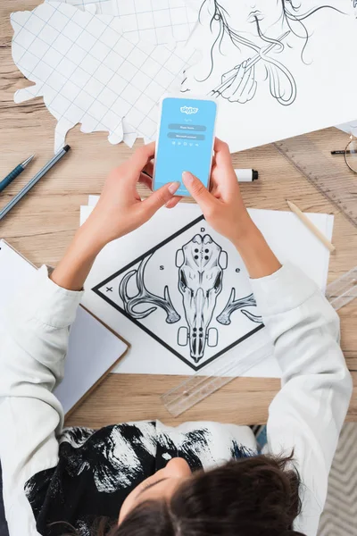 Overhead view of female designer using smartphone with skype application on screen at working table with paintings — Stock Photo