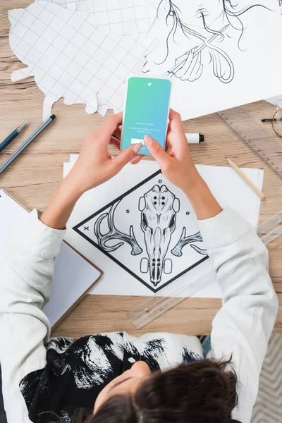 Overhead view of female designer using smartphone with twitter application on screen at working table with paintings — Stock Photo