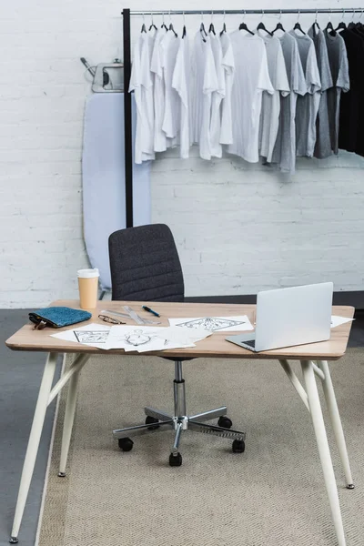 Foco seletivo de camisetas em cabides e mesa de trabalho com laptop e pinturas em estúdio de design de roupas — Fotografia de Stock