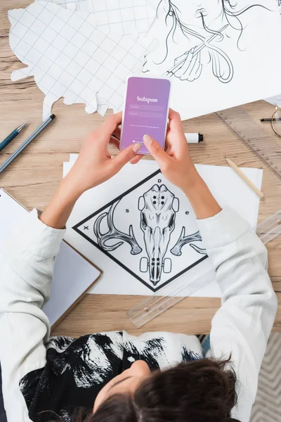 Overhead view of female designer using smartphone with instagram application on screen at working table with paintings — Stock Photo