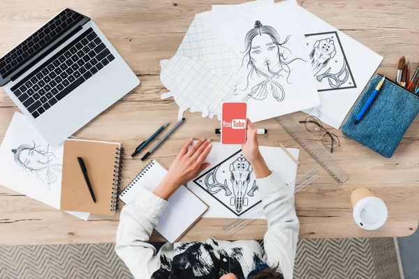 Vista aérea del diseñador femenino usando el teléfono inteligente con la aplicación de youtube en la pantalla en la mesa de trabajo con pinturas - foto de stock