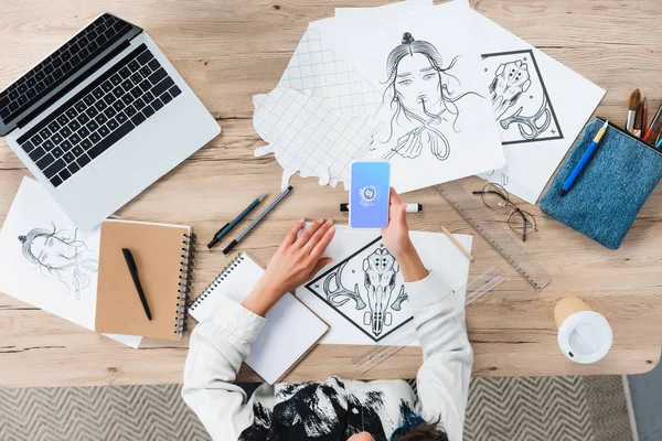 Overhead view of female designer using smartphone with shazam application on screen at working table with paintings — Stock Photo