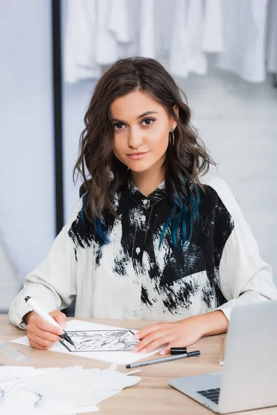 Sorridente pittura stilista femminile al tavolo di lavoro con computer portatile in studio di design di abbigliamento — Foto stock
