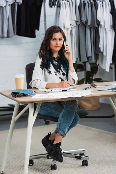 Sonriente joven diseñadora de moda hablando en smartphone en la mesa de trabajo en el estudio de diseño de ropa - foto de stock