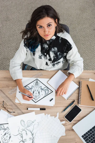 Vista aérea de la pintura artista femenina y mirando a la cámara mientras está sentado en la mesa con el ordenador portátil y el teléfono inteligente - foto de stock