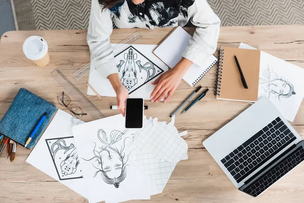 Imagen recortada del diseñador femenino sosteniendo teléfono inteligente con pantalla en blanco en la mesa de trabajo con pinturas y libros de texto - foto de stock