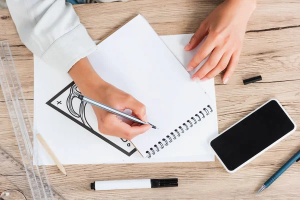 Cropped image of female fashion painting in textbook at table with smartphone — Stock Photo