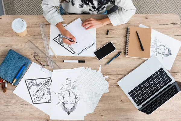 Обрезанное изображение женщины-художника, пишущей в учебнике за столом с ноутбуком и смартфоном — стоковое фото