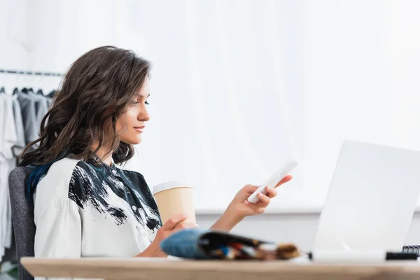 Sorridente designer de moda feminina com xícara de café usando smartphone à mesa com laptop — Fotografia de Stock