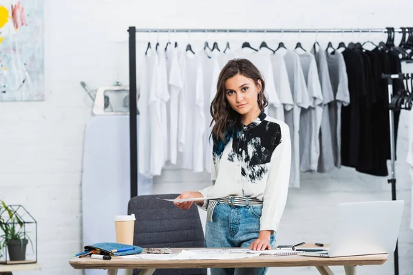 Attractive female fashion designer holding painting at table with laptop and coffee in clothing design studio — Stock Photo