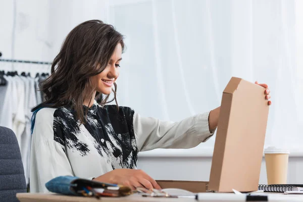 Sorrindo designer feminino abrindo caixa de pizza à mesa com xícara de papel de café — Fotografia de Stock