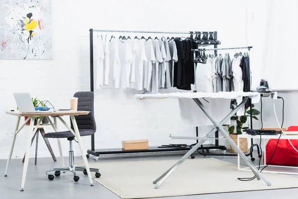 Selective focus of t-shirts on hangers, table with laptop and ironing board in clothing design studio — Stock Photo