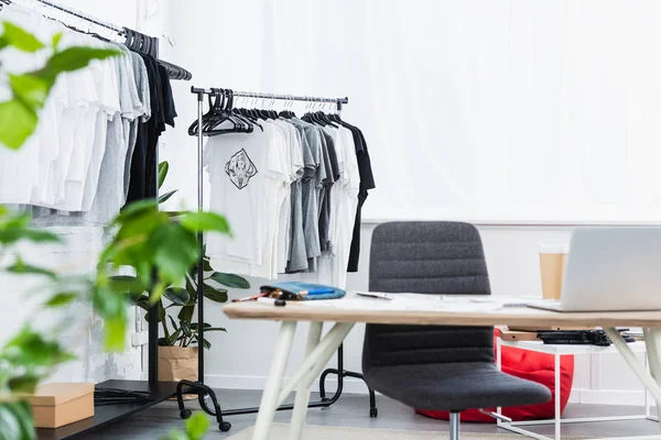 Enfoque selectivo de camisetas en perchas y mesa de trabajo con portátil en estudio de diseño de ropa - foto de stock