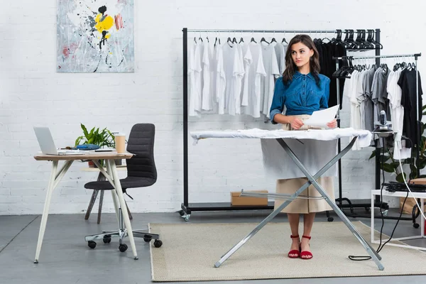 Diseñadora de moda femenina con pintura para imprimir en camiseta de pie cerca de tabla de planchar en estudio de diseño de ropa - foto de stock