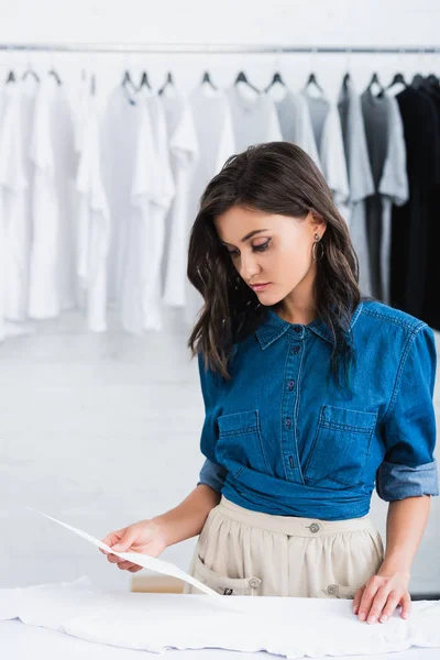 Joven y atractiva diseñadora de moda femenina eligiendo la impresión para la camiseta blanca vacía en el estudio de diseño de ropa - foto de stock