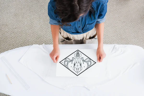 Vista aérea del diseñador de moda femenino que elige la impresión para la camiseta blanca vacía en la tabla de planchar - foto de stock