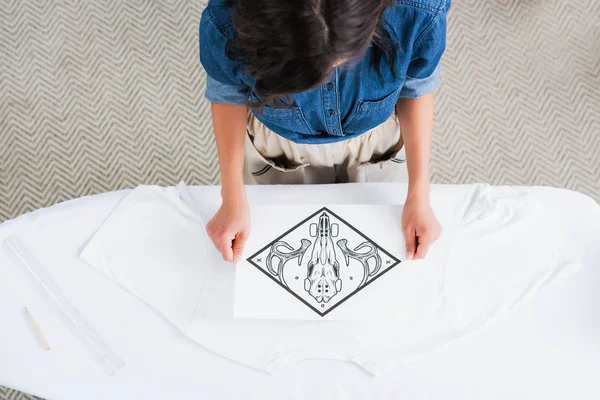 Overhead view of female fashion designer choosing print for empty white t-shirt at ironing board — Stock Photo