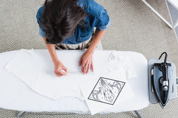 Overhead view of female fashion designer measuring space for print on empty white t-shirt at ironing board — Stock Photo