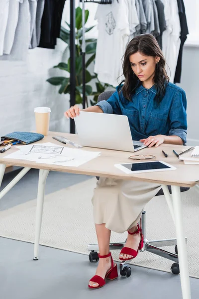 Серьезная женщина модельер работает за столом с ноутбуком в студии — стоковое фото