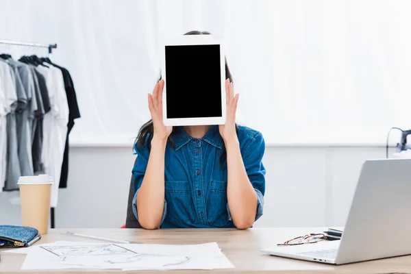 Mujer joven cubriendo cara por tableta digital con pantalla en blanco en la mesa de trabajo - foto de stock