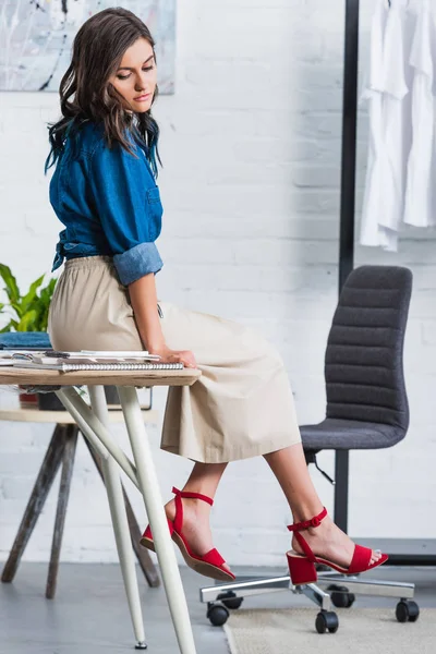 Atractiva diseñadora de moda sentada en la mesa en el estudio de diseño de ropa - foto de stock