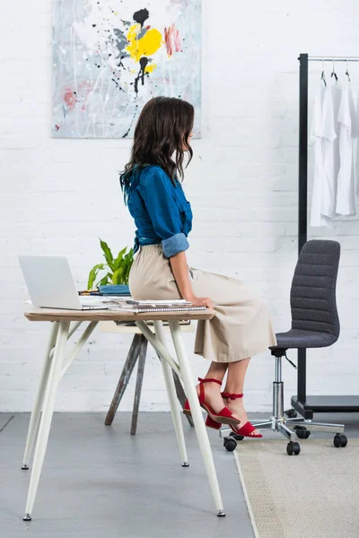 Side view of female fashion designer sitting on table in clothing design studio — Stock Photo